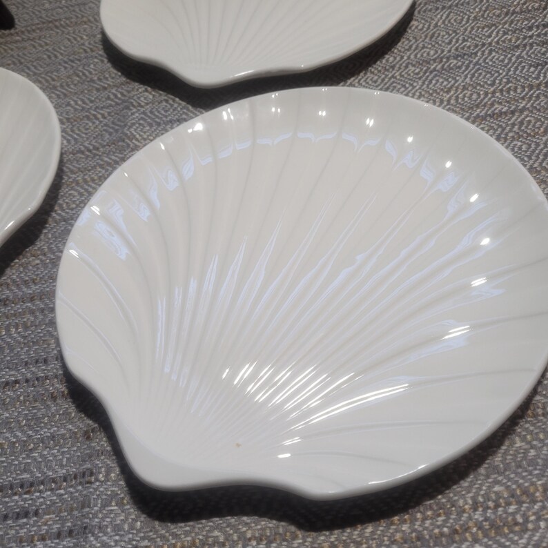 May include: A set of white ceramic plates shaped like scallop shells. Each plate features a glossy finish and raised, radiating ridges that mimic the natural texture of a seashell, extending from the base to the scalloped outer edge.