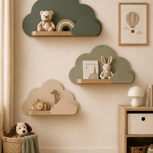 Peut inclure: Trois étagères murales en forme de nuage dans des tons vert sauge et beige. L'étagère supérieure porte un ours en peluche et un arc-en-ciel. L'étagère centrale présente un livre intitulé Kuçuk Prens et un lapin. L'étagère inférieure montre un appareil photo en bois et une girafe.
