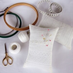 May include: Two white pincushions with embroidered designs, one with pink and gold accents, surrounded by sewing supplies including two embroidery hoops, white thread spools, and a pair of gold scissors.