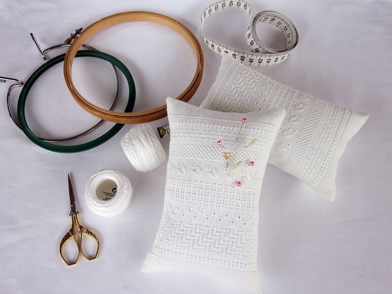 May include: Two white pincushions with embroidered designs, one with pink and gold accents, surrounded by sewing supplies including two embroidery hoops, white thread spools, and a pair of gold scissors.