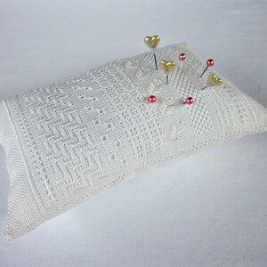May include: A white crocheted pincushion with gold heart-shaped pins and red glass beads.