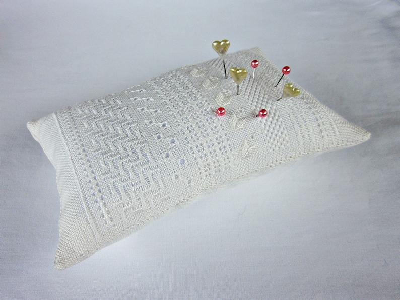 May include: A white crocheted pincushion with gold heart-shaped pins and red glass beads.
