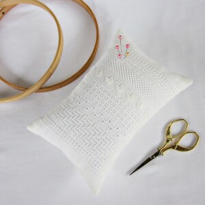 May include: White needlepoint pincushion with a delicate pattern and pink beads. The pincushion is sitting on a white surface with two wooden embroidery hoops and gold embroidery scissors.