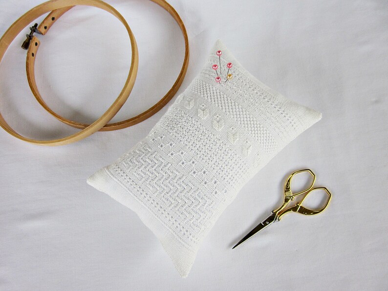 May include: White needlepoint pincushion with a delicate pattern and pink beads. The pincushion is sitting on a white surface with two wooden embroidery hoops and gold embroidery scissors.