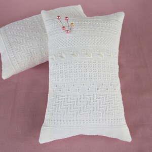 May include: Two white pincushions with intricate embroidered designs. The pincushion on the right has pink and gold pins inserted into it.
