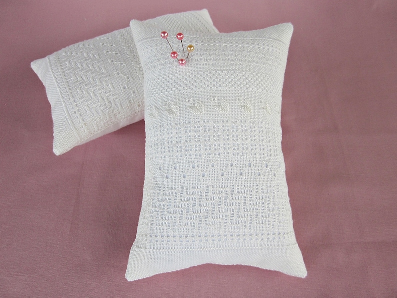 May include: Two white pincushions with intricate embroidered designs. The pincushion on the right has pink and gold pins inserted into it.