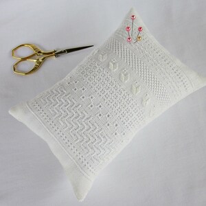 May include: White pincushion with a delicate lace pattern and pink beads. The pincushion is lying on a white surface with a pair of gold scissors.