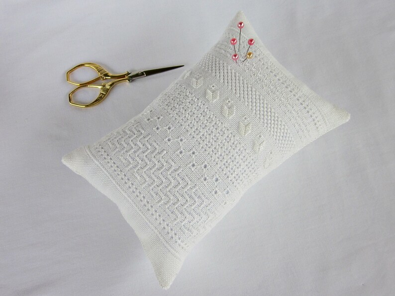 May include: White pincushion with a delicate lace pattern and pink beads. The pincushion is lying on a white surface with a pair of gold scissors.