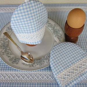 May include: Two blue and white gingham egg cozies with white lace trim. One cozy is on a plate with a spoon, the other is next to a wooden egg cup with a brown egg.