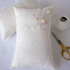 May include: Two white needlepoint pincushions with a delicate pattern, one with pink and white heart-shaped pins. A white spool of thread and gold scissors are also included.