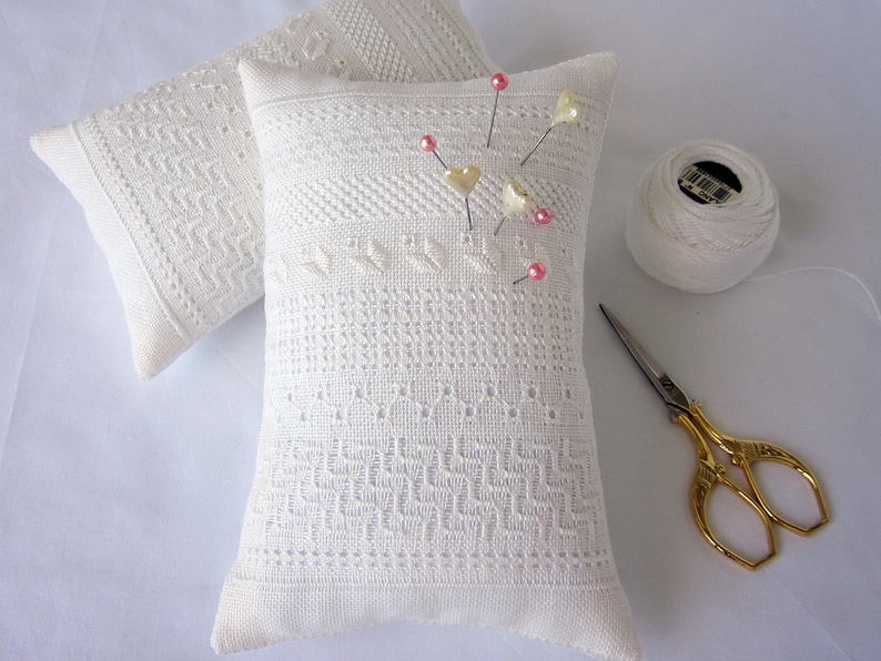 May include: Two white needlepoint pincushions with a delicate pattern, one with pink and white heart-shaped pins. A white spool of thread and gold scissors are also included.