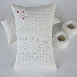 May include: Two white pincushions with delicate embroidery and a pin with red beads on top. The pincushions are on a white surface with two spools of white thread.