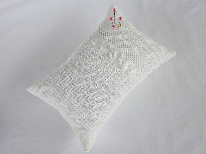 May include: White crocheted pillow with a delicate pattern and a small cluster of pink and gold beads pinned to the top.