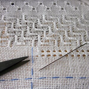 May include: Close-up of white fabric with a needle and thread, a pair of scissors, and blue lines marking the fabric. The fabric has a pattern of small holes and a delicate lace design.