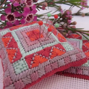 May include: A small, square, pink and green needlepoint pincushion with a geometric pattern. The pincushion is filled with pins with green and red heads.