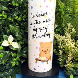 May include: A white cylindrical tumbler featuring a colorful pattern of small polka dots in blue, yellow, pink, and purple. The text "Cursive is the new Ig-pay Atin-lay" is printed in black script above a cartoon pig writing in a notebook at a desk.