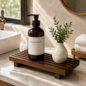 May include: A rectangular dark wood slatted tray with four short feet, holding a brown glass pump bottle with a white label reading HAND SOAP and a small, speckled off-white ceramic vase containing a green leafy plant sprig.