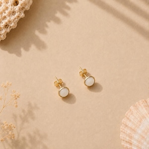 May include: A pair of gold stud earrings featuring a smooth, round, opaque white stone set in a polished gold metal bezel. The earrings include standard gold post backs and are displayed as a matching set on a neutral, light-colored surface.