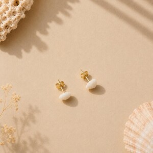 May include: A pair of gold-colored stud earrings with white, oval-shaped stones. The earrings are displayed on a beige surface, with a seashell and coral in the background. The earrings have a simple, elegant design.