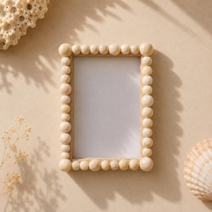 Op de afbeelding: Een rechthoekige houten fotolijst met een kralenrand. De lijst is leeg, met een wit interieur. De lijst is omgeven door schelpen en gedroogde planten, allemaal op een beige achtergrond. De lijst is ongeveer 20 cm bij 25 cm.