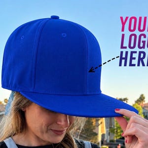Peut inclure: Casquette de baseball bleu vif avec une visière plate. La casquette présente une couronne structurée et un bouton bleu assorti. Le texte "YOUR LOGO HERE" est affiché sur le côté de la casquette.