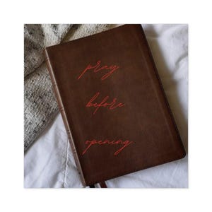 May include: A brown leather-bound book with the words "pray before opening" in red script. The book is lying on a white surface with a gray and white textured fabric in the background. A bookmark is visible.
