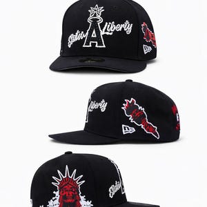 Op de afbeelding: Zwarte baseball cap met witte "Statue of Liberty" tekst en afbeeldingen. Voorzien van een rood-witte Statue of Liberty afbeelding aan de zijkant. De cap heeft een platte klep en een New Era logo.