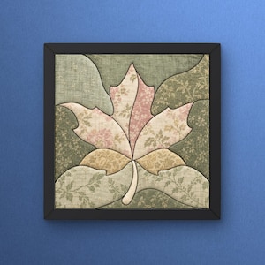 May include: A square framed wall art piece featuring a central maple leaf design composed of fabric patches. The leaf displays light pink and cream floral patterns, while the surrounding background consists of sage green fabric with leaf motifs.