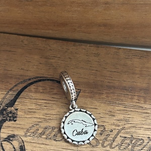 K&ouml;nnte beinhalten: Ein silberner Charm mit einem runden Anh&auml;nger mit gewelltem Rand. In der Mitte des Anh&auml;ngers befindet sich eine eingravierte Umrisskarte von Kuba &uuml;ber dem Wort Cuba in Schreibschrift. Der Charm hat eine &Ouml;se mit einem texturierten Perlenmuster.