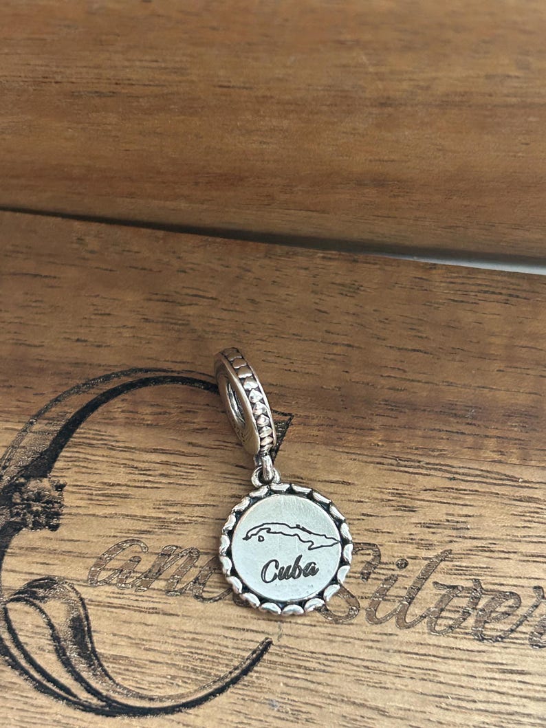 K&ouml;nnte beinhalten: Ein silberner Charm mit einem runden Anh&auml;nger mit gewelltem Rand. In der Mitte des Anh&auml;ngers befindet sich eine eingravierte Umrisskarte von Kuba &uuml;ber dem Wort Cuba in Schreibschrift. Der Charm hat eine &Ouml;se mit einem texturierten Perlenmuster.