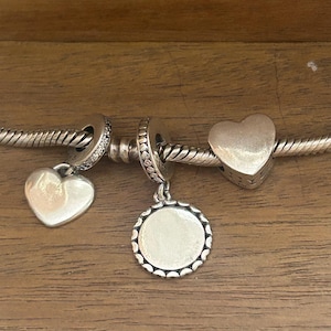 K&ouml;nnte beinhalten: Ein silbernes Bettelarmband mit drei verschiedenen Anh&auml;ngern: einem glatten herzf&ouml;rmigen Anh&auml;nger, einer runden Scheibe mit gewelltem Rand und einer massiven herzf&ouml;rmigen Perle. Die Armbandkette hat eine gedrehte, seilartige Struktur.