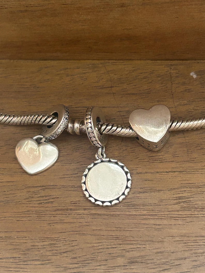 K&ouml;nnte beinhalten: Ein silbernes Bettelarmband mit drei verschiedenen Anh&auml;ngern: einem glatten herzf&ouml;rmigen Anh&auml;nger, einer runden Scheibe mit gewelltem Rand und einer massiven herzf&ouml;rmigen Perle. Die Armbandkette hat eine gedrehte, seilartige Struktur.