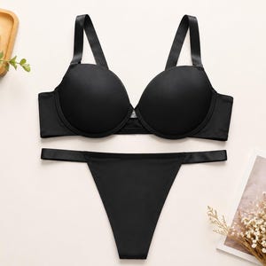 Könnte beinhalten: Ein schwarzes, passendes Lingerie-Set bestehend aus einem glatten Bügel-BH mit geformten Körbchen und verstellbaren Trägern sowie einem dazu passenden, hoch geschnittenen Tanga mit schmalem Bund, alles in einheitlichem, mattem Schwarz.