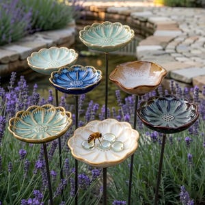 Può includere: Un set di sette picchetti da giardino in ceramica a forma di fiore su sottili aste metalliche. I petali presentano varie finiture smaltate nei toni dell'azzurro, blu scuro, marrone e bianco. Un fiore bianco ospita un'ape e sassolini di vetro.