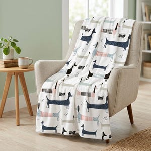 Op de afbeelding: Een witte plaid met een herhalend patroon van gestileerde teckels in marineblauw, lichtroze en grijstinten. De honden zijn afgebeeld met diverse texturen en het woord WOOF is in zwarte letters over het gehele ontwerp verspreid.