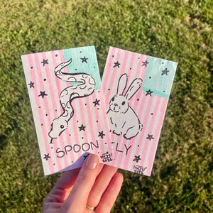 May include: Two rectangular cards featuring hand-drawn illustrations on a background of vertical pink stripes and a light blue rectangle. One card shows a snake, the other a rabbit, both surrounded by black stars. The text SPOONFLY is visible.