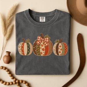 May include: A charcoal grey short-sleeve t-shirt featuring a graphic of three pumpkins with patchwork-style patterns in shades of orange, green, and brown. The centre pumpkin is adorned with a red and white checkered bow. The label reads Comfort Colors.