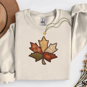 Peut inclure: Un sweat-shirt à col rond couleur sable arborant un motif central de feuille d'érable brodée. La feuille est composée de divers empiècements aux motifs écossais, vichy et floraux dans des tons automnaux, avec une petite citrouille orange.