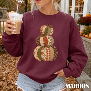 May include: A maroon long-sleeve sweatshirt featuring a central graphic of three stacked pumpkins. The pumpkins are decorated with a patchwork design of various autumn-themed patterns, including floral prints, plaid, and solid earth-tone colours.