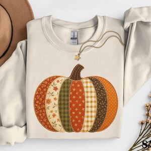 May include: A sand-coloured crewneck sweatshirt featuring a large, central pumpkin graphic composed of vertical fabric-style patches in various autumn-inspired patterns, including floral, gingham, and polka dot prints in shades of orange, green, and brown.