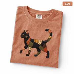 May include: A terracotta-colored t-shirt featuring a graphic of a black cat composed of various patchwork-style fabric patterns in shades of brown, orange, and cream. The shirt has a crew neckline and a Comfort Colors brand label at the inner neck.