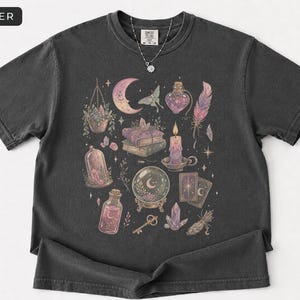 May include: A charcoal gray short-sleeve t-shirt featuring a graphic print of a crescent moon, moth, potion bottles, stacked books, a lit candle, a crystal ball, tarot cards, a key, crystals, a feather, and a hanging plant. The text PEPPER is visible.