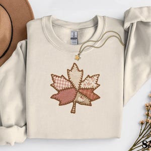 May include: A sand-colored crewneck sweatshirt featuring a central graphic of a large maple leaf composed of various patchwork fabric patterns in shades of pink, brown, and cream, with a small white pumpkin icon and a gold star necklace.