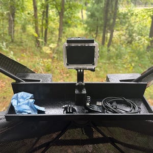 May include: A black rectangular tray mount for an action camera, featuring a central vertical stand with a mounted camera in a clear protective housing. The tray holds a light blue microfiber cloth, two black plastic mounting brackets, and a black USB cable.