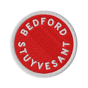 May include: Circular embroidered patch featuring a red central area with the white text "BEDFORD STUYVESANT". The patch has a white border. Suitable for sewing onto garments or bags, it's a decorative item.