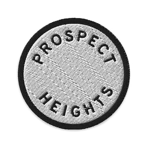 May include: A circular embroidered patch featuring a white center with the black text PROSPECT HEIGHTS arranged in a circular pattern along the border, finished with a thick black stitched edge that frames the entire design clearly.