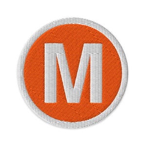 May include: Embroidered patch featuring a white letter "M" on a textured orange circle. The circle is bordered by a white stitched edge. The patch is circular and likely designed to be sewn or ironed onto fabric.