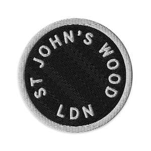 May include: A circular embroidered patch featuring a black center with the white text ST. JOHN'S WOOD and LDN arranged in a circular pattern. The patch has a thick, white stitched border that frames the black interior and the white lettering.