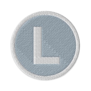 May include: A round, light blue patch with a white embroidered letter "L" in the center. The patch has a white border and a textured appearance. The design is simple and clean, suitable for various applications.