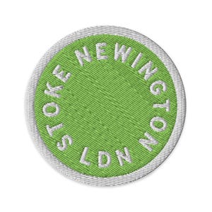 May include: A circular embroidered patch featuring a bright lime green center and a white border. The text STOKE NEWINGTON LDN is stitched in white capital letters in a circular pattern around the inner edge of the patch.